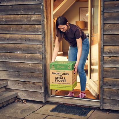 Woman carrying a box of short logs for small stoves outside a wooden building.