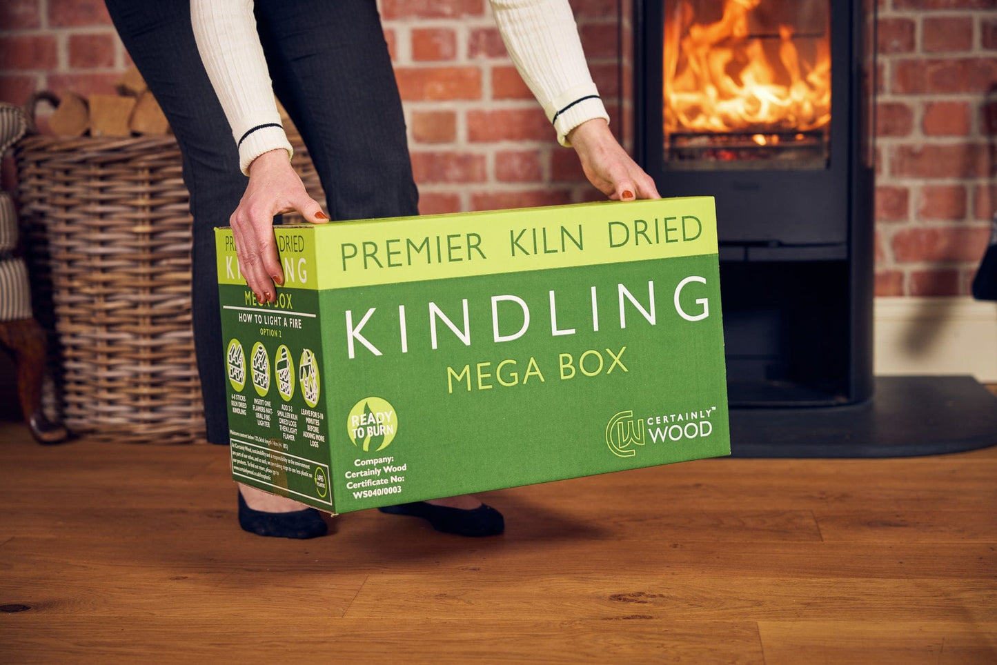 Person holding a green box of 'Premier Kiln Dried Kindling Mega Box' in front of a fireplace.