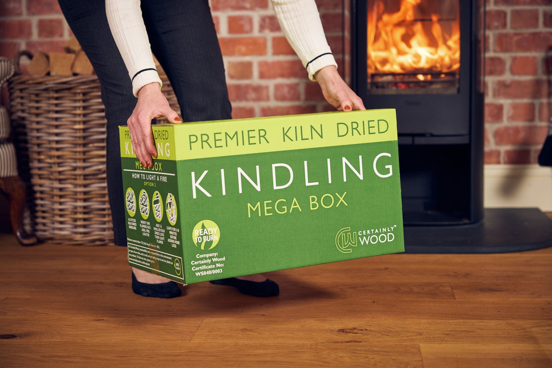 Person holding a green box of 'Premier Kiln Dried Kindling Mega Box' in front of a fireplace.