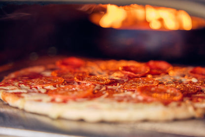 Pepperoni pizza baking in a wood-fired oven with flames.